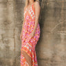 Full body front view of model wearing the Trip To The Tropics Maxi Dress that has orange, fuchsia, purple, pink and ivory fabric with a floral pattern,  two tiered body, a halter neck, a cinched waist, and an open back.