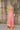 Full body back view of model wearing the Trip To The Tropics Maxi Dress that has orange, fuchsia, purple, pink and ivory fabric with a floral pattern,  two tiered body, a halter neck, a cinched waist, and an open back.