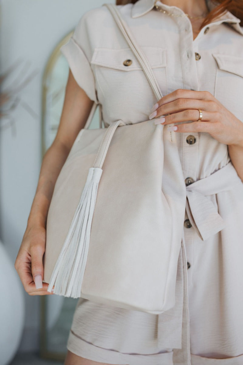 Close up view of model wearing the Getaway Plan Purse which features ivory faux leather fabric, snap button closure, fringe details and two skinny shoulder straps.