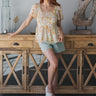 Full body front view of model wearing the See You In My Dreams Top that has white sheer fabric with a floral pattern, white lining, a smocked chest and back, a square neck, short sleeves, and peplum hem.