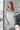Front view of model wearing the All Eyes On Me Dress that has white sheen fabric, white lining, mini length, an overlapped hem, one shoulder strap, and a monochromatic back zipper.