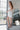 Full body side view of model wearing the All Eyes On Me Dress that has white sheen fabric, white lining, mini length, an overlapped hem, one shoulder strap, and a monochromatic back zipper.