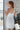 Back view of model wearing the All Eyes On Me Dress that has white sheen fabric, white lining, mini length, an overlapped hem, one shoulder strap, and a monochromatic back zipper.