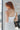 Upper back view of model wearing the All Eyes On Me Dress that has white sheen fabric, white lining, mini length, an overlapped hem, one shoulder strap, and a monochromatic back zipper.