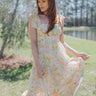 Full body view of model wearing the Boardwalk Sunset Dress which features white sheer fabric with a yellow, green, orange, and pink floral pattern, white lining, a maxi-length hem, a three-tiered skirt, a square neckline, tie straps, and a smocked back.