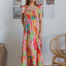 Full body view of model wearing the Destination Trip Dress which features pink, orange, yellow, green and white fabric, floral pattern, maxi dress, tiered body style, smocked chest, ruffle sweetheart neckline and tie straps.