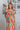 Front view of model wearing the Destination Trip Dress which features pink, orange, yellow, green and white fabric, floral pattern, maxi dress, tiered body style, smocked chest, ruffle sweetheart neckline and tie straps.