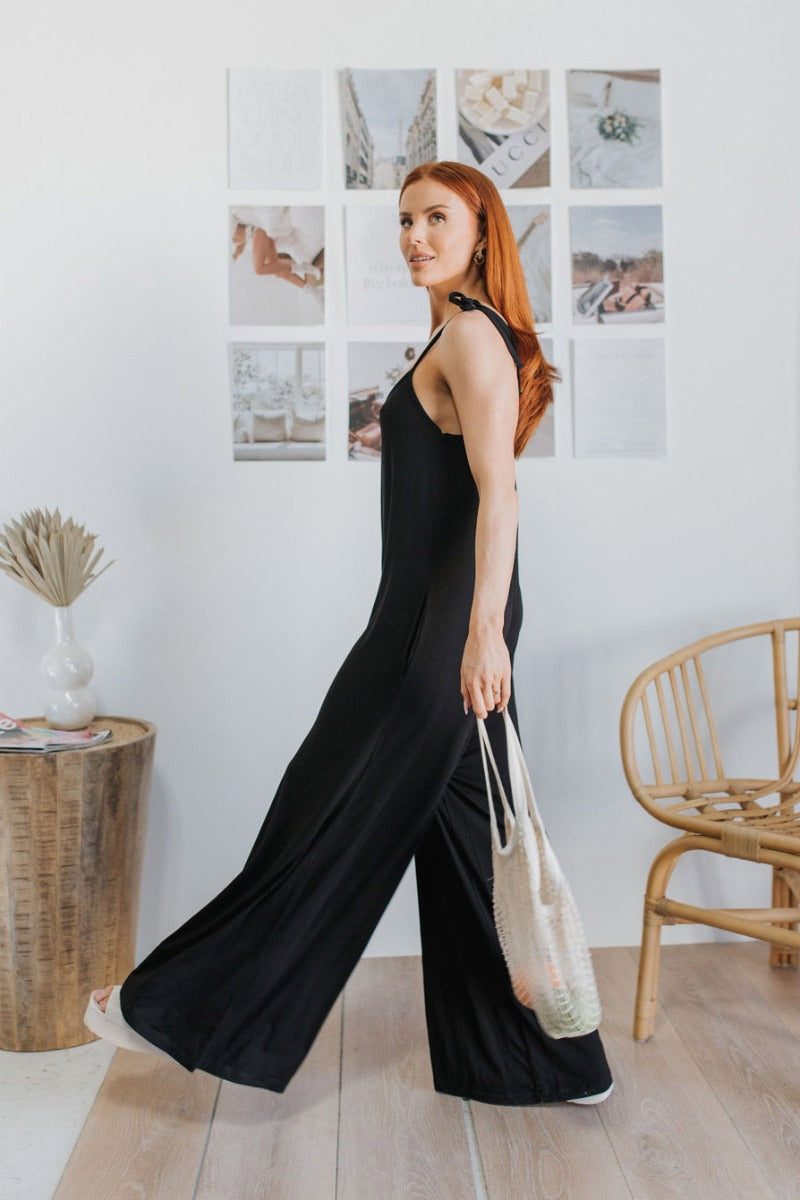 Side view of model wearing the Stepping Out Jumpsuit which features black fabric, v-neckline, spaghetti strap ties and long flare pants.