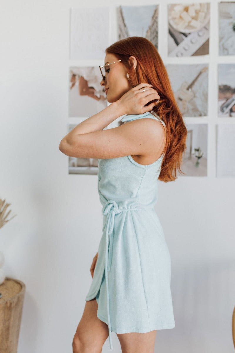 Side view of model wearing the Sky Views Romper which features light blue terry cloth fabric, an overlapped hem, a surplice neckline, a tie around the waist for closure, a collared neckline, and a sleeveless design.