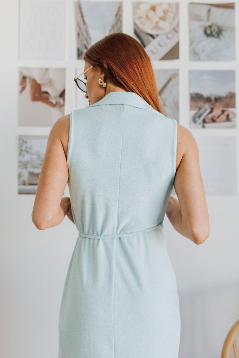Back view of model wearing the Sky Views Romper which features light blue terry cloth fabric, an overlapped hem, a surplice neckline, a tie around the waist for closure, a collared neckline, and a sleeveless design.