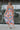 Full body back view of model wearing the Pacific Coast Floral Midi Dress that has purple, orange, pink, green, blue and cream fabric, floral and palm leaf print, and adjustable straps.