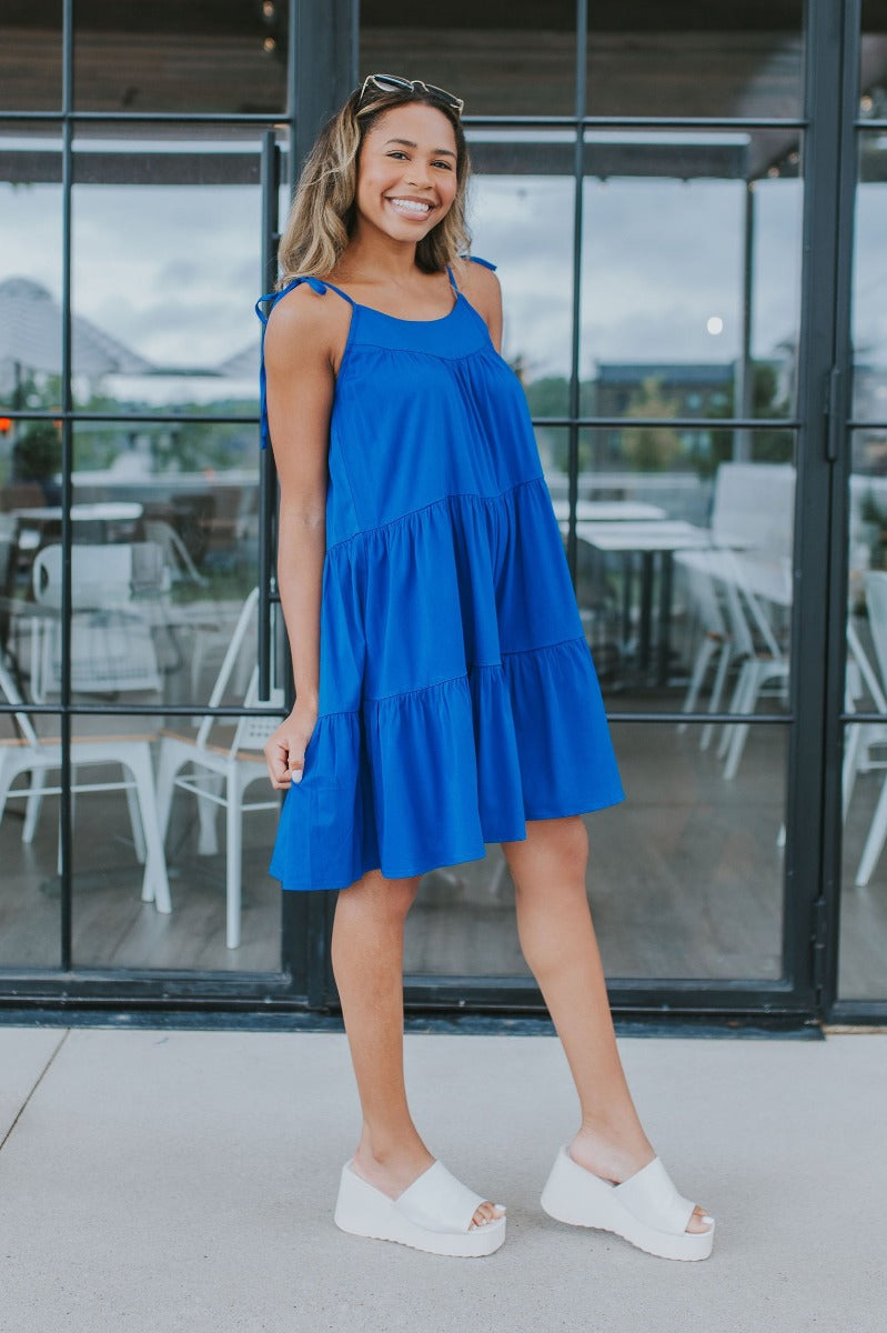 Full body side view of model wearing the Summer Shade Romper that has cobalt blue fabric, a two-tiered body, a scooped neckline, and tie straps.