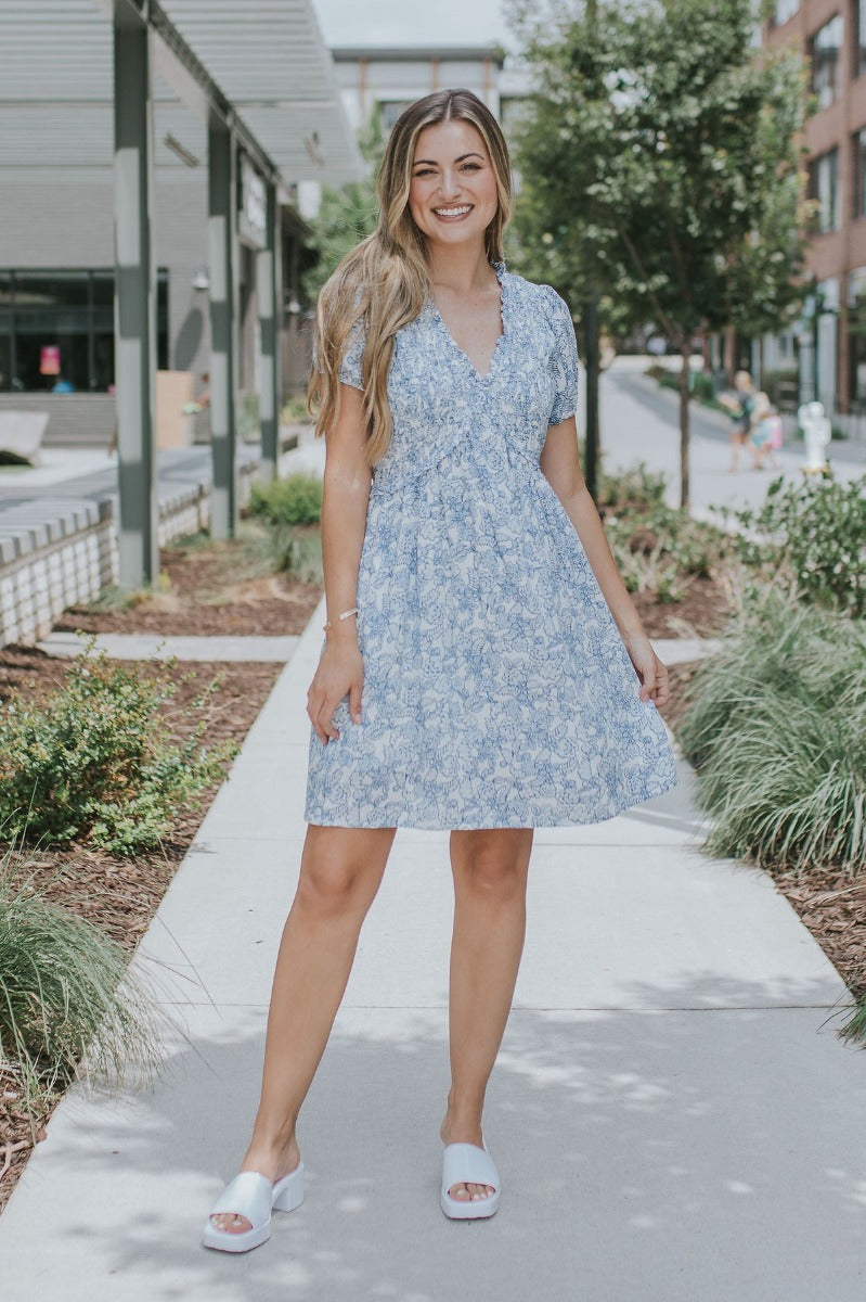 Full body front view of model wearing the Ocean Boulevard Floral Dress that has white fabric with blue florals, white lining, a v-neck, a smocked upper, an elastic waist, a back keyhole, and puff sleeves
