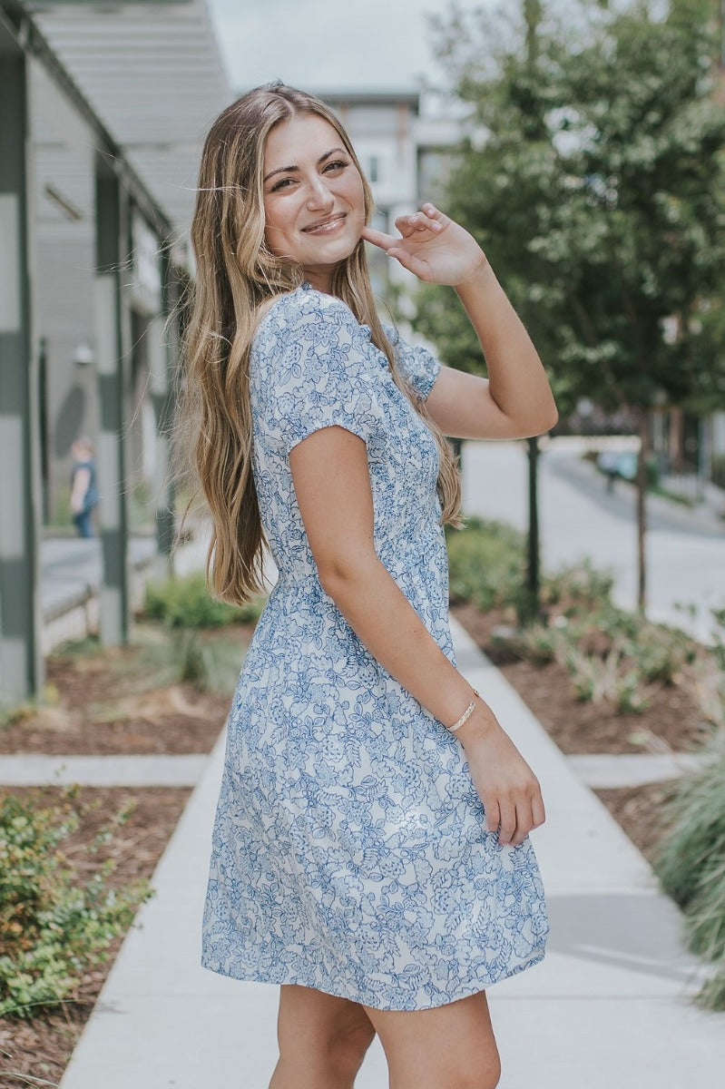 side view of model wearing the Ocean Boulevard Floral Dress that has white fabric with blue florals, white lining, a v-neck, a smocked upper, an elastic waist, a back keyhole, and puff sleeves