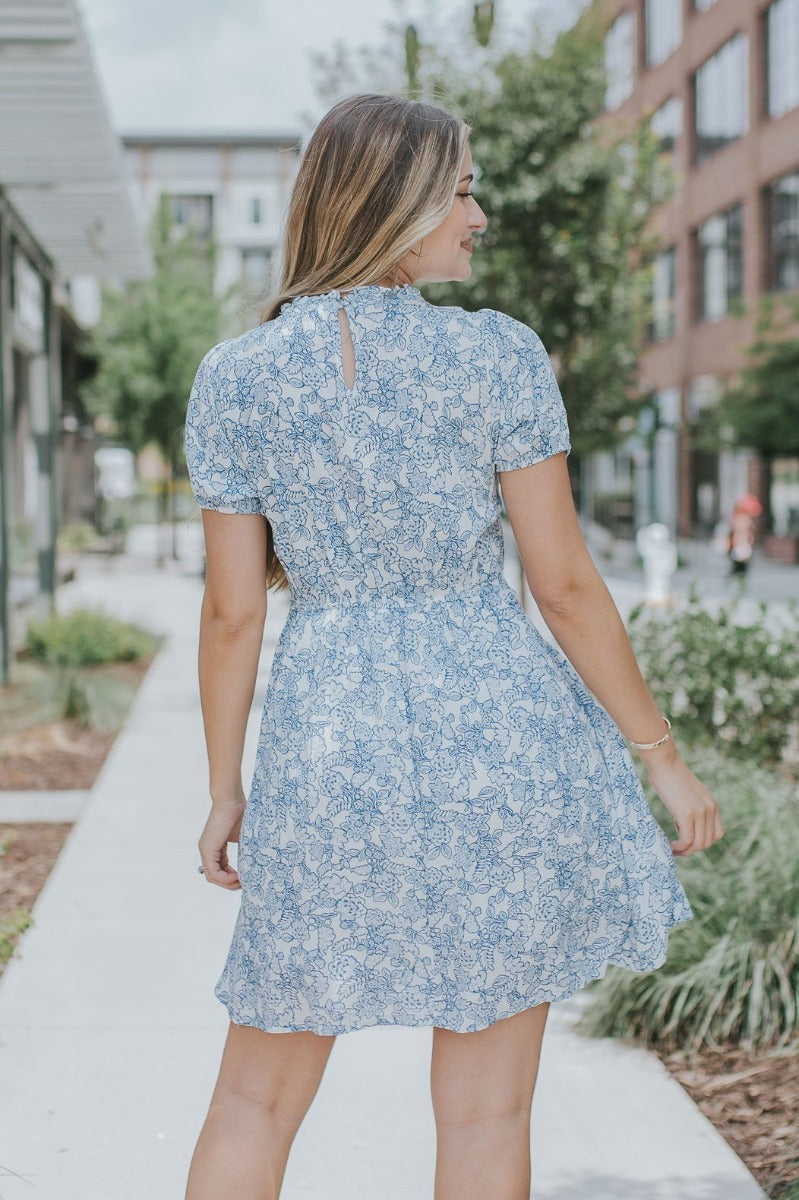 back view of model wearing the Ocean Boulevard Floral Dress that has white fabric with blue florals, white lining, a v-neck, a smocked upper, an elastic waist, a back keyhole, and puff sleeves