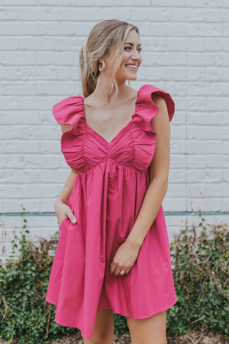 Front view of model wearing the Total Knockout Dress in Pink which features magenta fabric, mini length, a v-neckline, side pockets, pleated upper details, dramatic ruffle straps, magenta lining, a smocked upper, and a monochromatic back zipper with a hoo