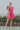 Full body view of model wearing the Total Knockout Dress in Pink which features magenta fabric, mini length, a v-neckline, side pockets, pleated upper details, dramatic ruffle straps, magenta lining, a smocked upper, and a monochromatic back zipper with a