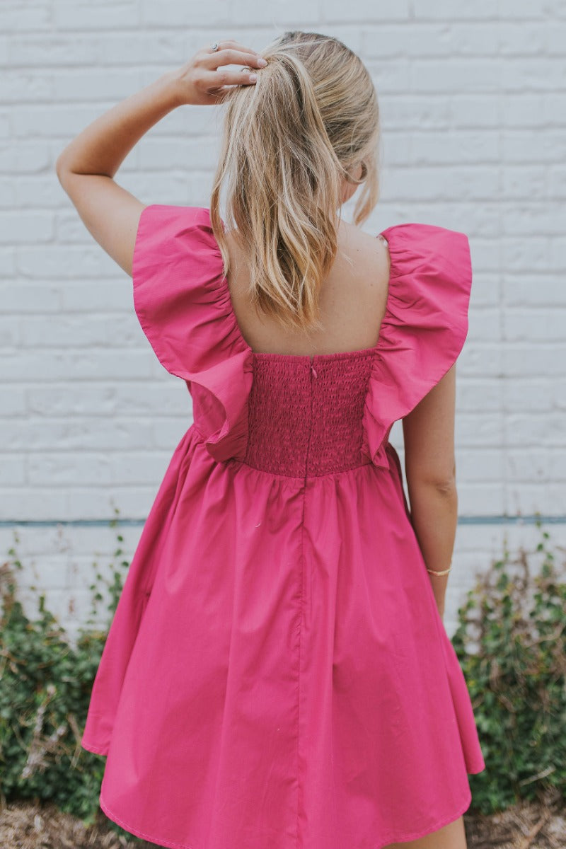 Back view of model wearing the Total Knockout Dress in Pink which features magenta fabric, mini length, a v-neckline, side pockets, pleated upper details, dramatic ruffle straps, magenta lining, a smocked upper, and a monochromatic back zipper with a hook