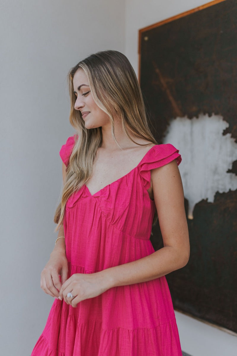 Side view of model wearing the Sweet Energy Mini Dress which features hot pink fabric, a mini hem length, a three-tiered body, hot pink lining, a v-neckline, and ruffle straps.