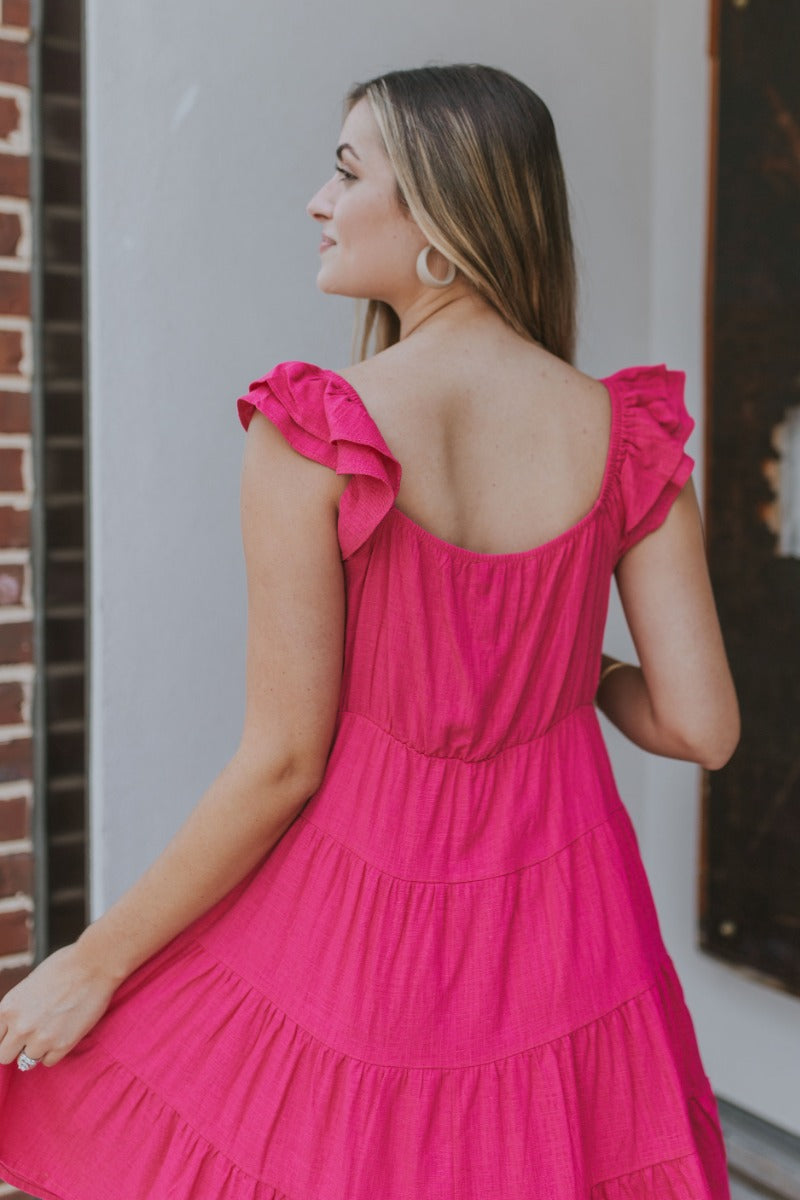 Back  view of model wearing the Sweet Energy Mini Dress which features hot pink fabric, a mini hem length, a three-tiered body, hot pink lining, a v-neckline, and ruffle straps.