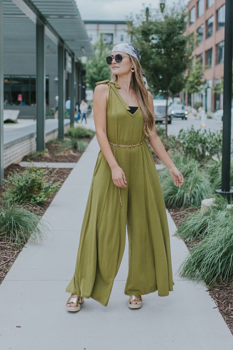Full body view of model wearing the Fall For You Jumpsuit which features olive green knit fabric, pockets on each side, a v-neckline, tie straps, and flared pant legs.