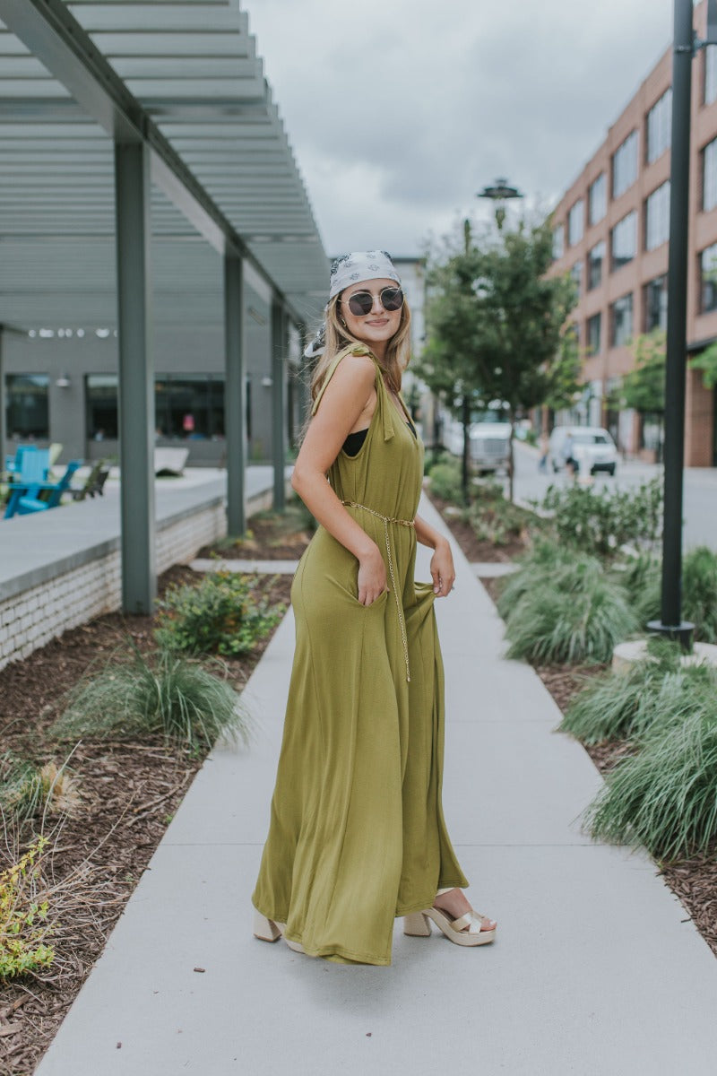 Side view of model wearing the Fall For You Jumpsuit which features olive green knit fabric, pockets on each side, a v-neckline, tie straps, and flared pant legs.