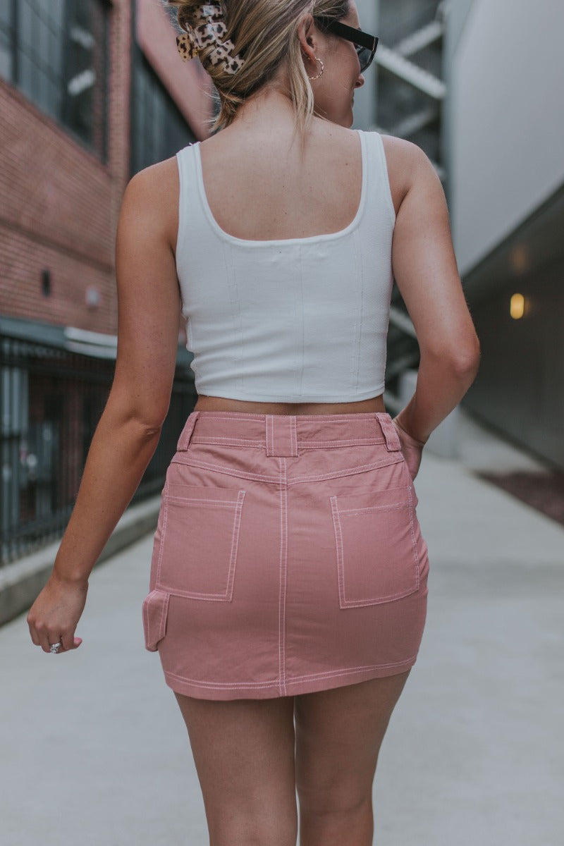 Back view of model wearing the Back To Back Skirt which features mauve fabric, mini length, two front pockets, two back pockets, white contrast stitching, a front zipper with double button closures, and belt loops.
