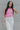 Front view of model wearing the Speak Your Mind Bodysuit which features pink fabric, round neckline, sleeveless and thong bottom with button clasp closures.