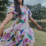 Front view of model wearing the Color Your World Midi Dress that has white fabric with watercolor spots,  a three tiered body, a square neck, a smocked back, and short bubble sleeves.