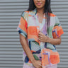 Front view of model wearing the West Coast Top which features orange, purple, yellow, blue, green and white sheer fabric, geometric water color pattern, purple button down for closure, collared neckline and short sleeves.