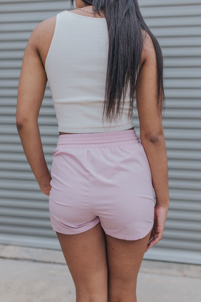 Back view of model wearing the Casually Chic Shorts which features light pink fabric, two side pockets with covered button closure, elastic waistband and drawstring tie.