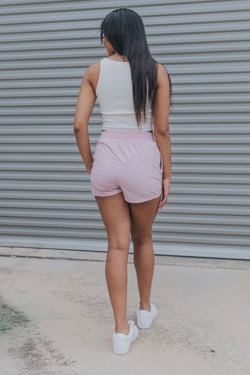 Full body back view of model wearing the Casually Chic Shorts which features light pink fabric, two side pockets with covered button closure, elastic waistband and drawstring tie.