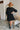 Back view of the Comfort Zone Dress In Black that features a black colored material, a round neck with a ruffle trim, a V neckline with an adjustable tie closure, a 3/4 sleeve, a tiered design, and a mini length.