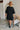 Back view of the Comfort Zone Dress In Black that features a black colored material, a round neck with a ruffle trim, a V neckline with an adjustable tie closure, a 3/4 sleeve, a tiered design, and a mini length.