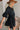Side view of the Comfort Zone Dress In Black that features a black colored material, a round neck with a ruffle trim, a V neckline with an adjustable tie closure, a 3/4 sleeve, a tiered design, and a mini length.