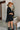 Side view of the Comfort Zone Dress In Black that features a black colored material, a round neck with a ruffle trim, a V neckline with an adjustable tie closure, a 3/4 sleeve, a tiered design, and a mini length.