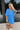 Frontal view of the Comfort Zone Dress that features a blue colored material, a round neck with a ruffle trim, a V neckline with an adjustable tie closure, a 3/4 sleeve, a tiered design, and a mini length.