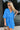 Frontal view of the Comfort Zone Dress that features a blue colored material, a round neck with a ruffle trim, a V neckline with an adjustable tie closure, a 3/4 sleeve, a tiered design, and a mini length.