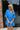 Frontal view of the Comfort Zone Dress that features a blue colored material, a round neck with a ruffle trim, a V neckline with an adjustable tie closure, a 3/4 sleeve, a tiered design, and a mini length.