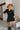 Frontal view of the Comfort Zone Dress In Black that features a black colored material, a round neck with a ruffle trim, a V neckline with an adjustable tie closure, a 3/4 sleeve, a tiered design, and a mini length.