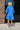 Back view of the Comfort Zone Dress that features a blue colored material, a round neck with a ruffle trim, a V neckline with an adjustable tie closure, a 3/4 sleeve, a tiered design, and a mini length.