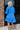 Back view of the Comfort Zone Dress that features a blue colored material, a round neck with a ruffle trim, a V neckline with an adjustable tie closure, a 3/4 sleeve, a tiered design, and a mini length.