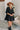 Frontal view of the Comfort Zone Dress In Black that features a black colored material, a round neck with a ruffle trim, a V neckline with an adjustable tie closure, a 3/4 sleeve, a tiered design, and a mini length.