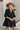 Frontal view of the Comfort Zone Dress In Black that features a black colored material, a round neck with a ruffle trim, a V neckline with an adjustable tie closure, a 3/4 sleeve, a tiered design, and a mini length.