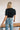 Back view of the Cool and Classy Top that features a black colored material, a round neckline with a lace trim, a half sleeve, a smocked body, a ruffle trim, and a back button closure.