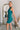 Side view of the Tell Me About It Dress that features a teal ribbed material, a high round neckline, a sleeveless design with one shoulder thick strap, a tight fit, and a mini length.