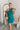 Frontal view of the Tell Me About It Dress that features a teal ribbed material, a high round neckline, a sleeveless design with one shoulder thick strap, a tight fit, and a mini length.