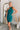 Frontal view of the Tell Me About It Dress that features a teal ribbed material, a high round neckline, a sleeveless design with one shoulder thick strap, a tight fit, and a mini length.