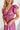 Frontal view of the Living In A Dream Maxi Dress that features a pink colored material, a V neckline, a short ruffle sleeve, a knot detail at the bust, an open back with adjustable tie closures, and a maxi length.