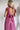 Back view of the Living In A Dream Maxi Dress that features a pink colored material, a V neckline, a short ruffle sleeve, a knot detail at the bust, an open back with adjustable tie closures, and a maxi length.